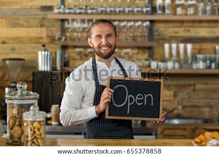 Portrait of smiling waiter standing with open sign board in cafe