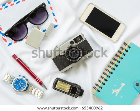 Flat Lay photo with props are Air letter, watch, pen, Flash, Camera, Glasses, Charger, Blue Notebook and smartphone on white fabric background.
