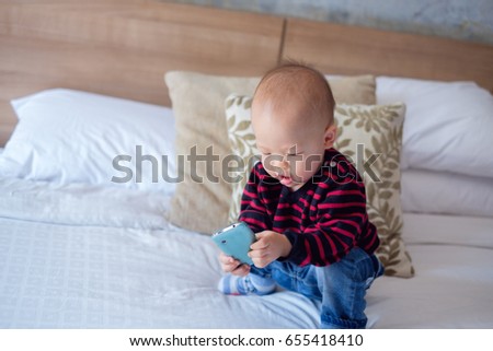 Cute asian toddler baby boy sitting in bed and holding & looking at smartphone. Kids playing in bedroom at home. leisure & children & technology & internet addiction concept - Selective focus