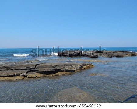 Lava slab rock along Argaka shoreline, Cyprus showing more slabs just under the surface of sunlit water in forefront  of picture. Blue sky and dark blue sea in background