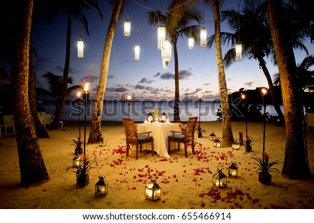 A Table Set up for a romantic meal on the beach with lanterns and chairs and flowers with palms and sky and sea in the background Royalty-Free Stock Photo #655466914