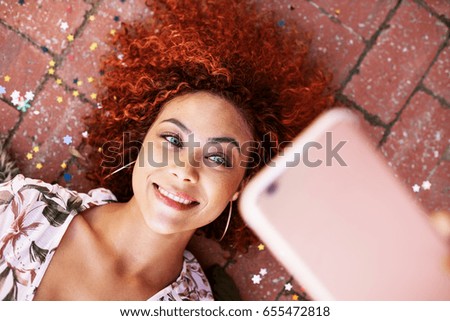 Close up of smiling woman in curly brown hair lying on floor looking at a smartphone. Woman posing for a selfie lying on the ground.