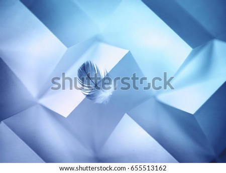 Abstract geometric blue background with triangles and rectangles of paper and a Light air feather in balance on the edge of the paper edge.