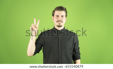 Young handsome man with black shirt makes victory sign. Isolated green background.
