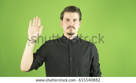 Young man with black shirt makes swearing sign with his hand open and up. Green background, isolated.