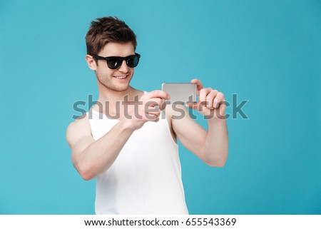 Picture of young happy man in sunglasses standing isolated over blue background using phone. Looking aside.
