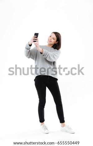 Full length portrait of a smiling pretty girl standing and taking selfie on mobile phone isolated over white background