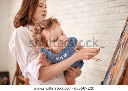 Portrait of young woman painting picture in art studio with curious baby girl in arms