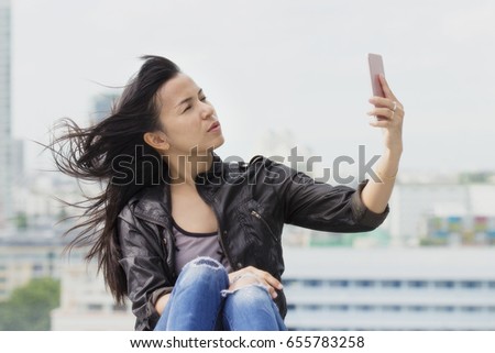 Attractive and confident woman holding smartphones to selfie over deck against windy on cityscape background.