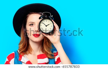 Portrait of young smiling red-haired white european woman in hat and red striped shirt with alarm clock on blue background