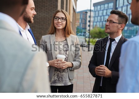 Multi-ethnic group of colleagues standing at office building and chatting animatedly with each other while having coffee break, over shoulder view