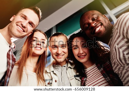 Low angle view of joyful friends posing for photography while standing against panoramic windows, waist-up group portrait