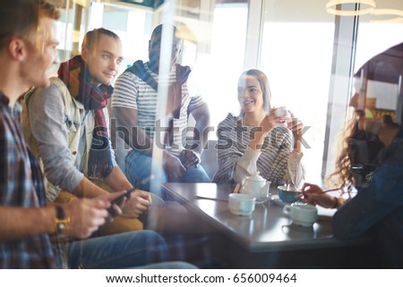 Multi-ethnic group of friends gathered together in coffeehouse and chatting animatedly with each other, view through glass wall