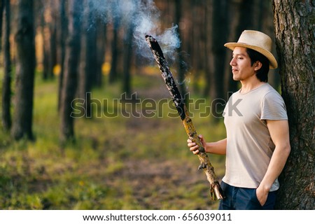 Native american indian peruvian indigenous man in straw hat standing above tree in forest with burning and smoking wooden stick. Shaman rite. Mystic ceremony. Fire  invocation. Old reservation