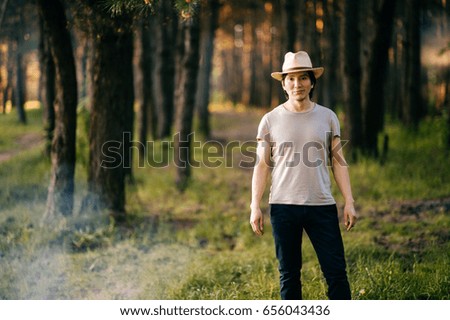Young and lonely native american indian latino indigenous man standing in forest outdoor in summer in straw hat, casual clothing above bonfire with smoke on vacation. Evening sunset. Weekend.