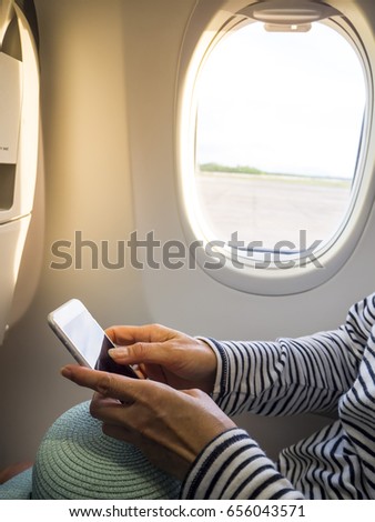 Woman passenger on airplane sitting window seat in casual t-shirt use smart phone.