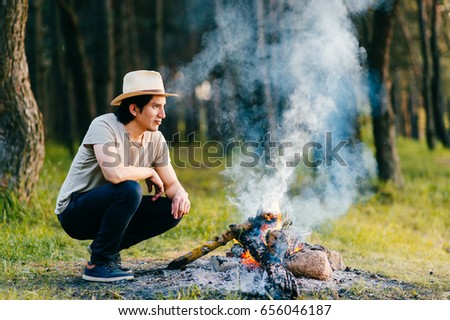 Native american indian peruvian indigenous man in straw hat sitting in forest outdoor in summer above bonfire with smoke and torch at hand. Shaman rite. Spiritual ceremony. Young sorcerer.
