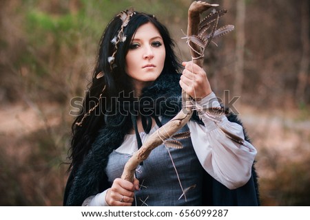 A beautiful girl with long black hair. Witch in a medieval dress and a black cloak with a pagan stick. Fantasy picture to Halloween. Model in the forest. Feathers in plaits, shamanic rite.