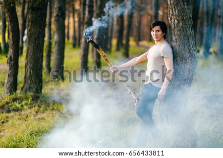 indigenous man standing with burning, smoking wooden stick.