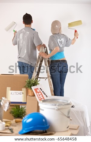 Couple standing on one ladder, painting wall together with paint roller.?