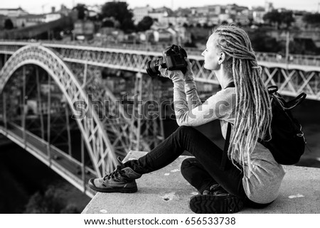 Young beautiful woman with dreadlocks and a camera sits taking pictures of a Dom Luis I bridge  in old Porto, Portugal. Tourism and travel, black and white photo.