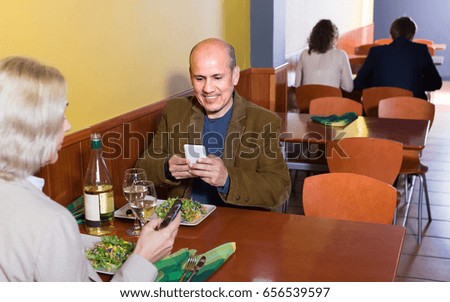 Smiling aged couple taking photos of salad with smartphones in restaurant 
