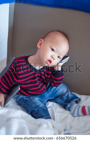 Cute asian toddler baby boy sitting in crib and talking on phone & holding tablet, pc Kids playing in baby cot. leisure & children & technology & internet addiction concept