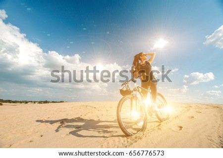Strong woman wearing in black sport suite sitting on bicycle and seeing on the sun. Fitness concept. Mountain bike rides along desert road in dunes. Background blue sky with clouds. 