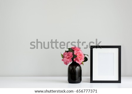 Pink camellia flowers in a black vase next to an empty black photo frame, on a white desktop. Horizontal with space for copy.