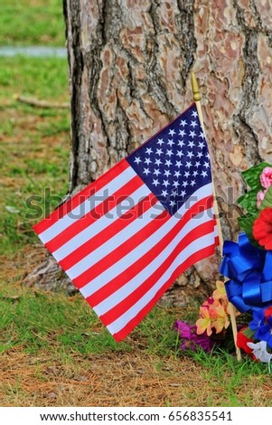 A US Flag flying at a Cemetery on Memorial  day with a tree in the back ground, with flower's and green grass,.