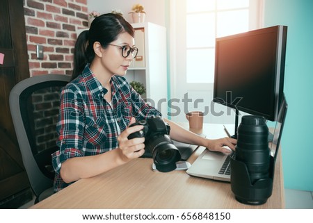 professional lovely female photographer holding business camera and using mobile laptop computer transport picture sitting on editing office desk.
