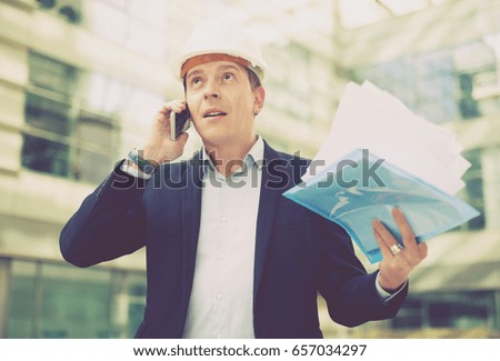 Smiling architect in helmet with paper documents in the hand talking phone