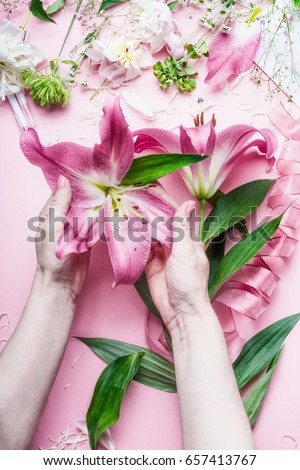 Creative Florist workspace. Female hands holding beautiful big pink lily flowers on pastell table with florist decoration equipment, top view, frame.Festive holiday concept