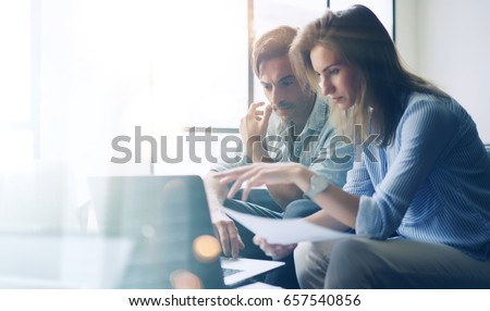Two young coworkers working on laptop computer at sunny office.Woman holding paper documents and pointing on notebook screen. Horizontal.Blurred background