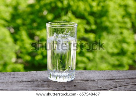 Glass with ice water on a wooden table against a background of green foliage