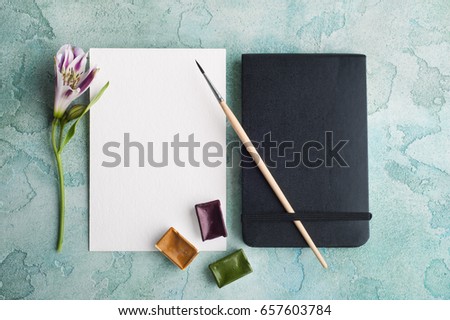 Paint brush, purple, yellow and green watercolor paint with spring flowers and blank white paper on blue concrete background.