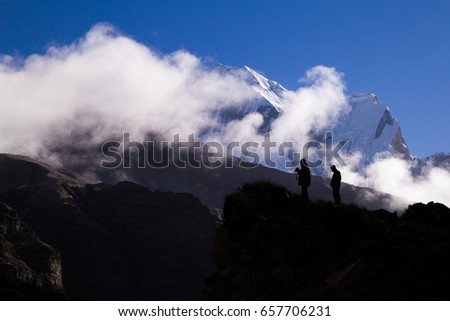 Morning view in high mountains. Silhouettes of two trekkers at the background of clouds and snow covered mountain peaks.