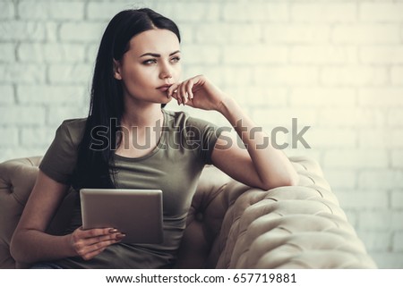 Beautiful pensive girl in casual wear is using a digital tablet and looking away while sitting on couch at home