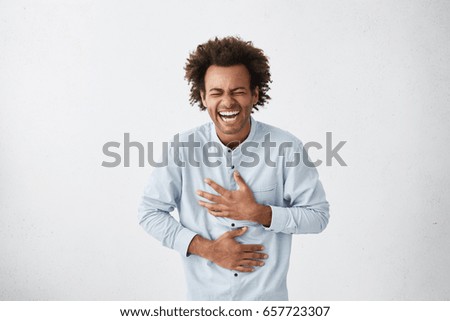 Positive and joyful Afro American male with fine crop of hair bursting into laughing holding his hands on stomach can`t stopping laughing after hearing funny anecdote. Positive emotions and humour Royalty-Free Stock Photo #657723307
