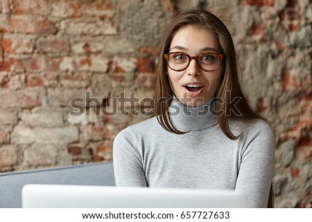 Pretty girl with surprised look having beautiful appearance and long straight hair wearing turtle neck and glasses sitting at cozy sofa using laptop for communication isolated over brick wall