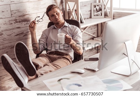 Handsome young pensive businessman is drinking coffee and resting in his chair in office