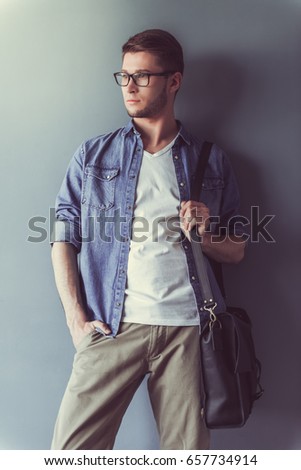 Handsome young businessman in casual clothes, glasses and with a stylish bag is looking away while leaning on the wall, on gray background