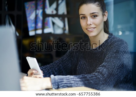 Portrait of charming young hipster girl sitting in coworking space looking at camera enjoying free time chatting in social networks via smartphone, attractive female sending messages using application