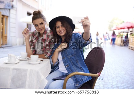 Two charming female dressed in trendy clothing making selfie and updating profile image in social network.Good looking young woman spending free time in sidewalk cafe and taking photo on smartphone
