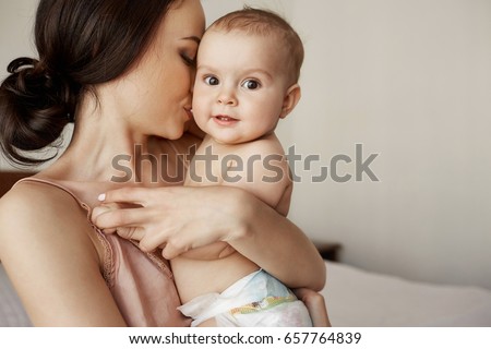 Young tender happy mother hugging her newborn baby smiling sitting on bed in morning.