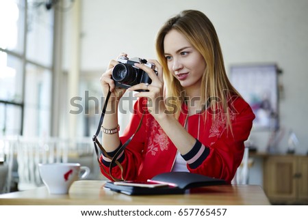 Charming attractive blonde female taking pictures on vintage camera sitting in coffee shop preparing poject, positive concentrated hipster girl making amateour pictures of cafe interior design