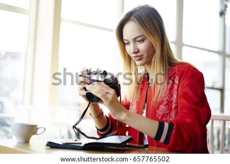 Attractive professional female photographer dressed in red trendy jacket making settings in vintage camera preparing old equipment for work,skilled hipster girl making amateur pictures sitting in cafe