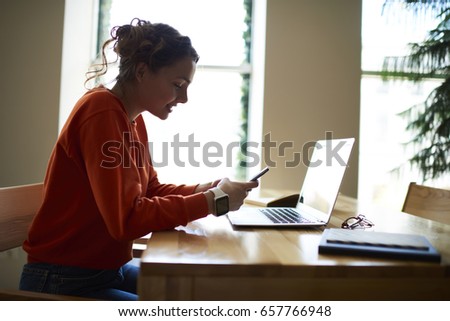 Smiling teenager browsing internet websites and doing shopping online on digital smartphone sitting at laptop and books in college interior.Young woman in good mood chatting with friends via cellular