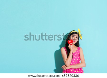 pretty sweet female student standing in blue wall background looking at empty area and holding heart daydreaming thinking future with cute dress.