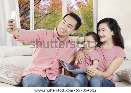 Portrait of happy family taking selfie picture by using smartphone while sitting on the sofa with autumn background on the window